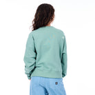Person wearing a light green sweatshirt with a logo on the back, standing against a white background.